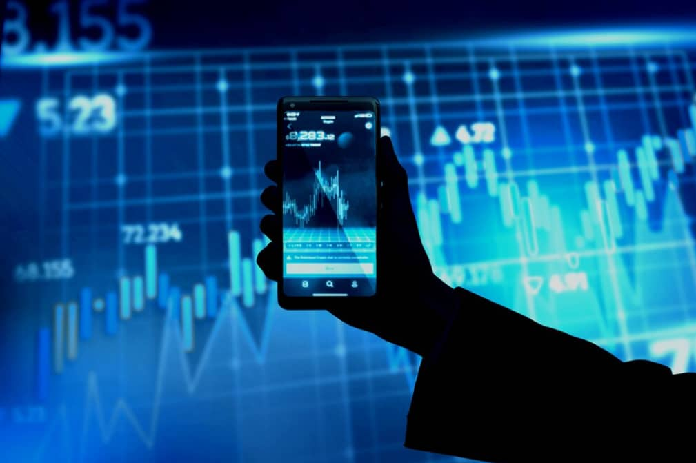 A hand holding a smartphone against a backdrop of stock market graphs