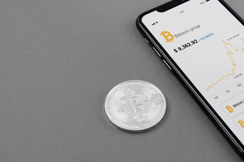 A silver Bitcoin coin beside a smartphone showing price charts